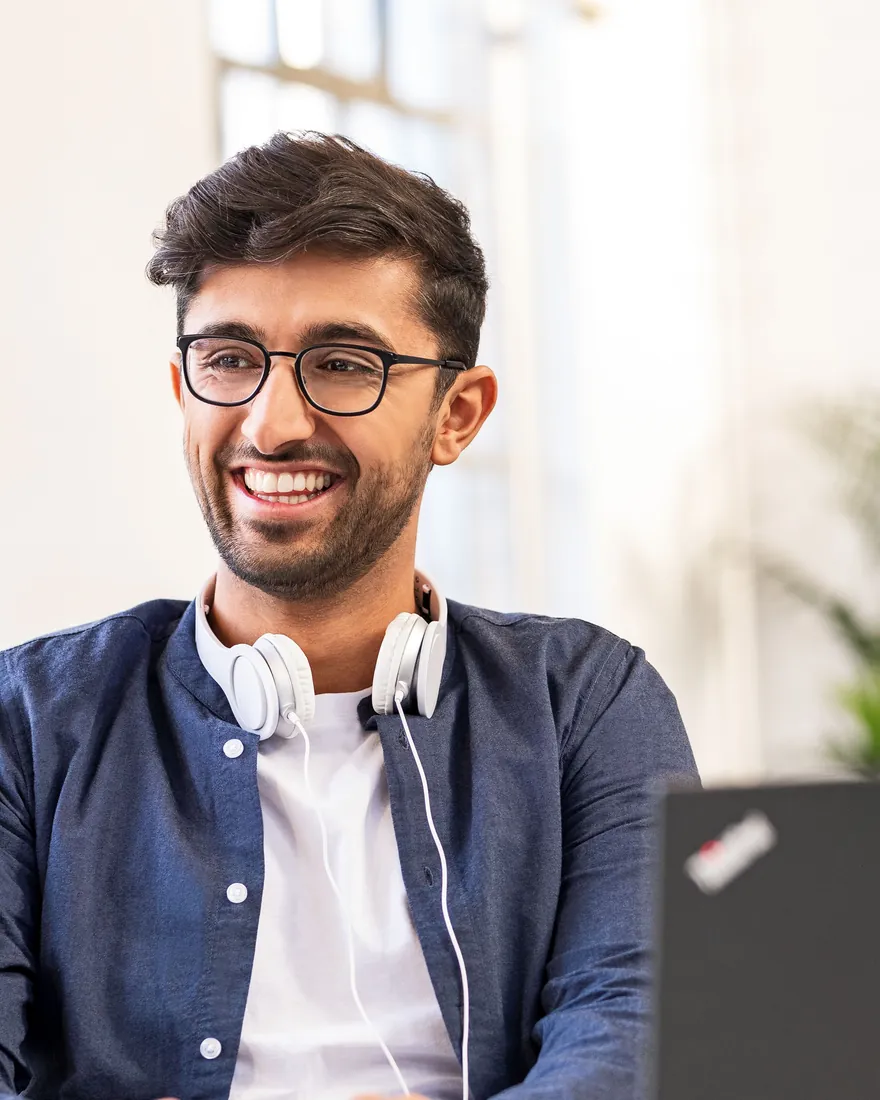Colaborador sorrindo de fones no pescoço, trabalhando no computador no escritório da Henkel em Düsseldorf
