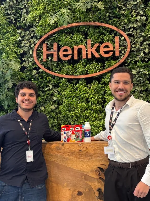 Dois estagiários sorrindo em frente ao painel verde com o logo da Henkel e produtos da marca