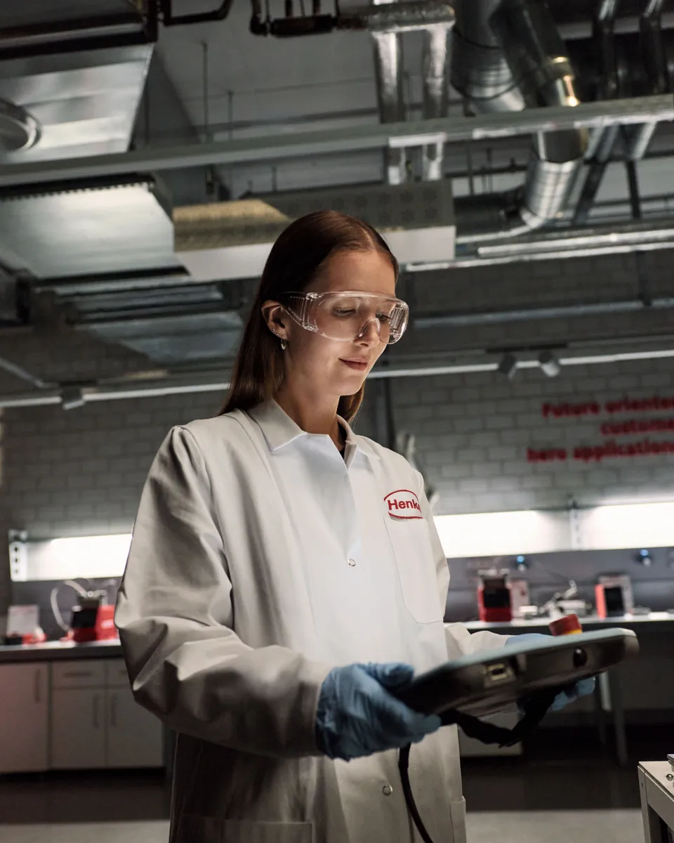 Pesquisadora de jaleco e óculos de proteção consultando um tablet no laboratório da Henkel